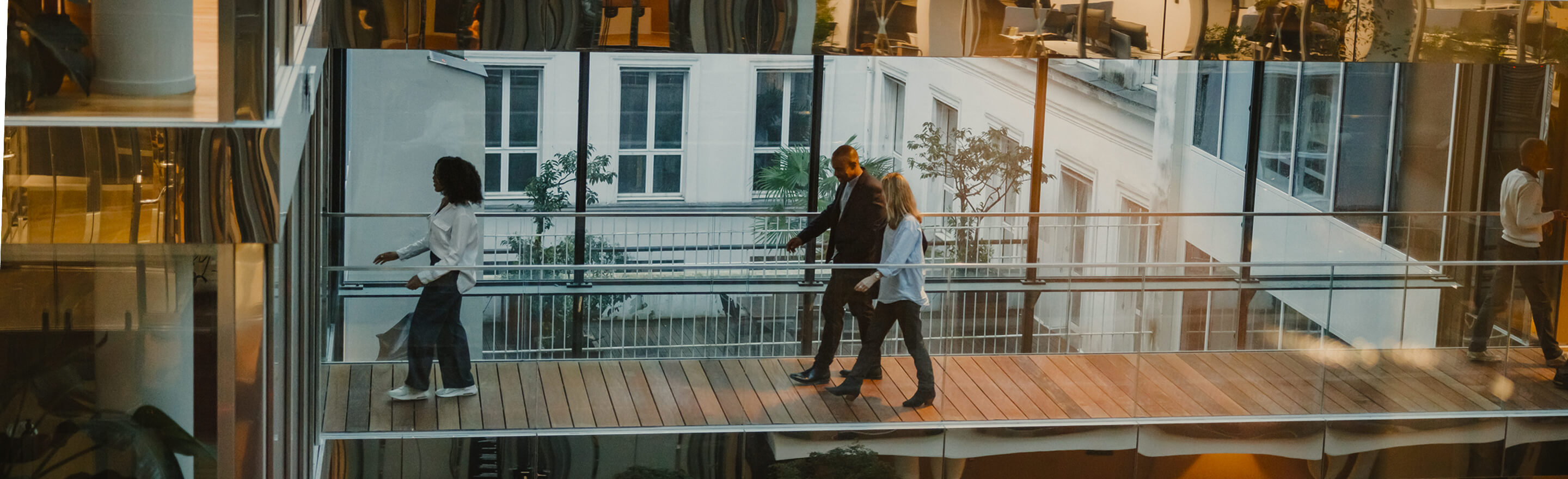 Men and women of various ethnic backgrounds walk and converse in a sleek, multi-level office, dressed in smart casual attire, surrounded by modern architecture and greenery.